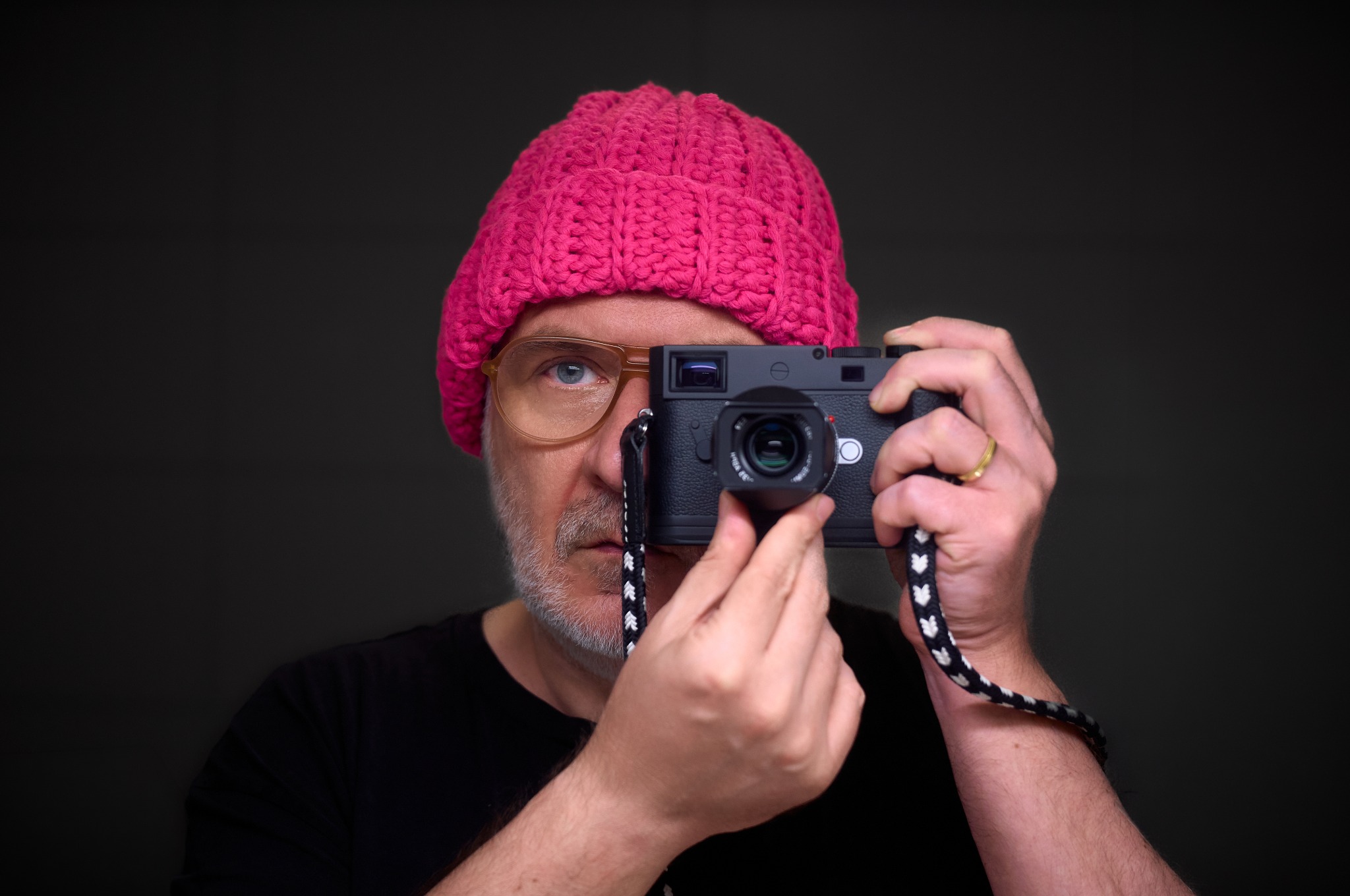 Ein Mann mit pinker Strickmütze und Brille hält eine Kamera vor sein Gesicht.